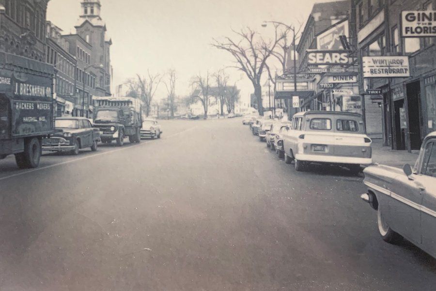 Image of Main Street in Haverhill, Massachusetts in the early 1900s.  This is evident by the types of vehicles parked along the road and the types of businesses and signs.  A company truck is shown in traffic.