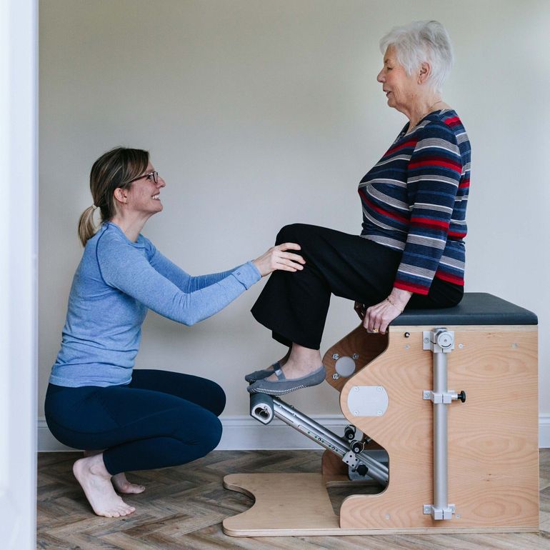 A woman is kneeling down next to a woman sitting on a pilates chair.