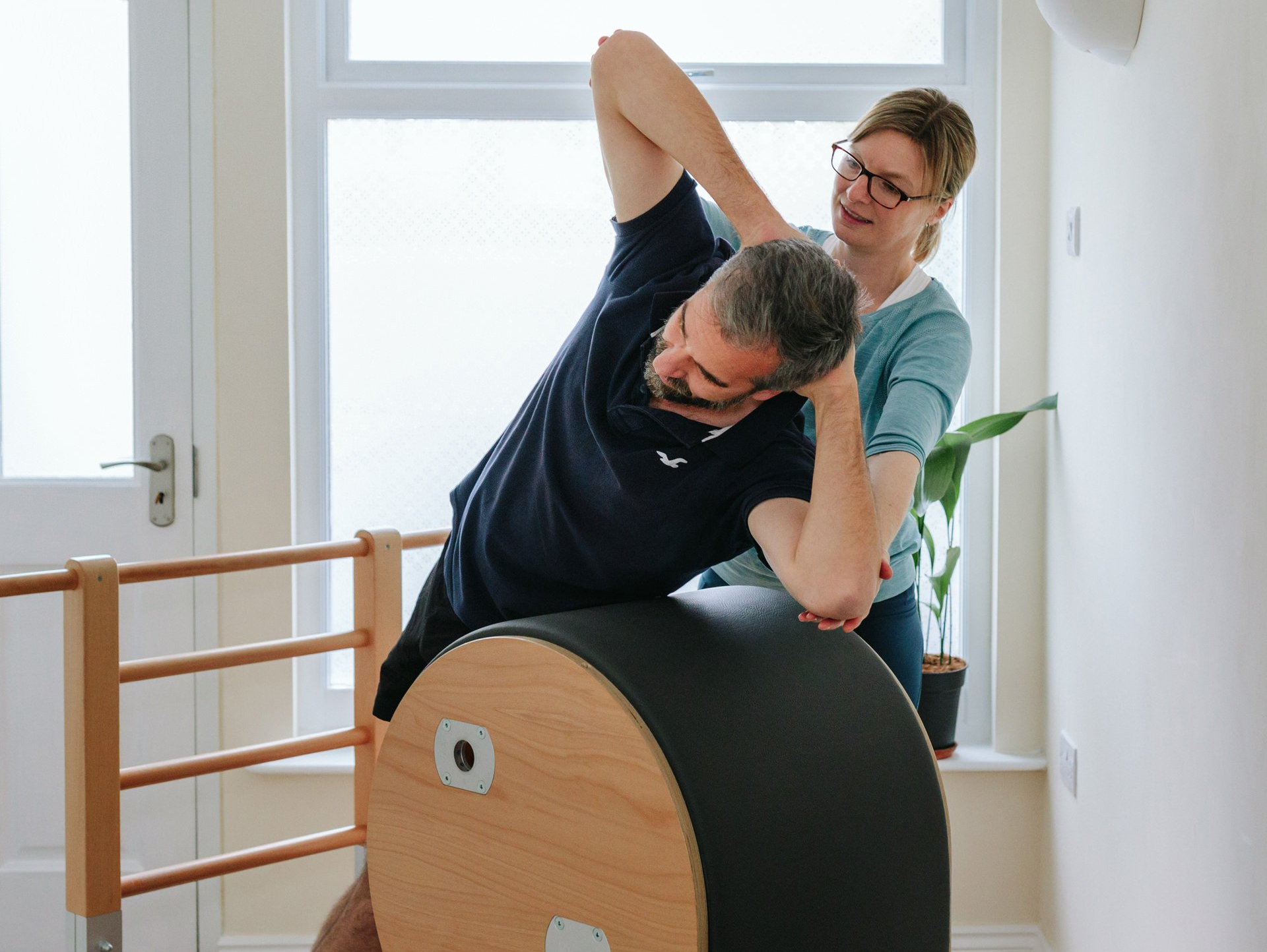 A woman is helping a man stretch his arms on a pilates barrel.