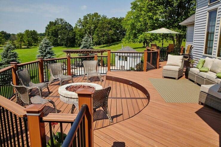 Spacious wooden deck with fire pit, seating areas, and a view of green fields under a sunny sky.