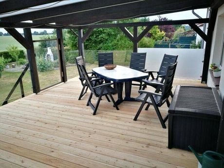 Wooden deck with a table and chairs under a dark wooden pergola, overlooking a garden.