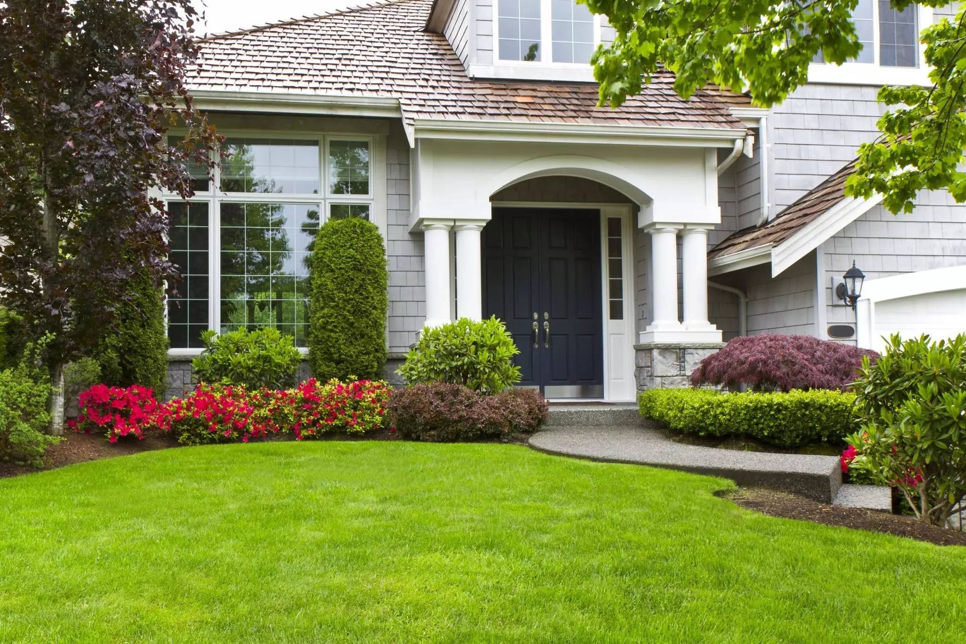 Gray house with a blue door, white columns, and lush green lawn and colorful flowerbeds.