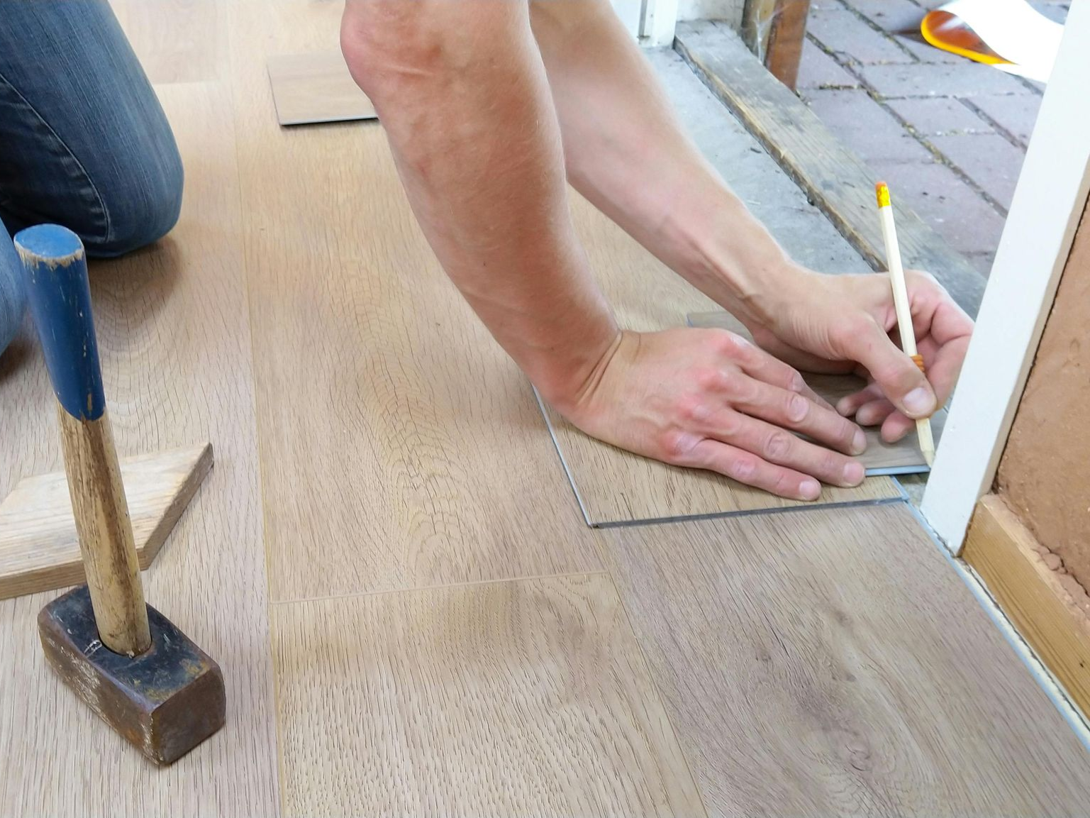 Person installing wood flooring, using pencil to mark a plank against door frame. Hammer nearby.
