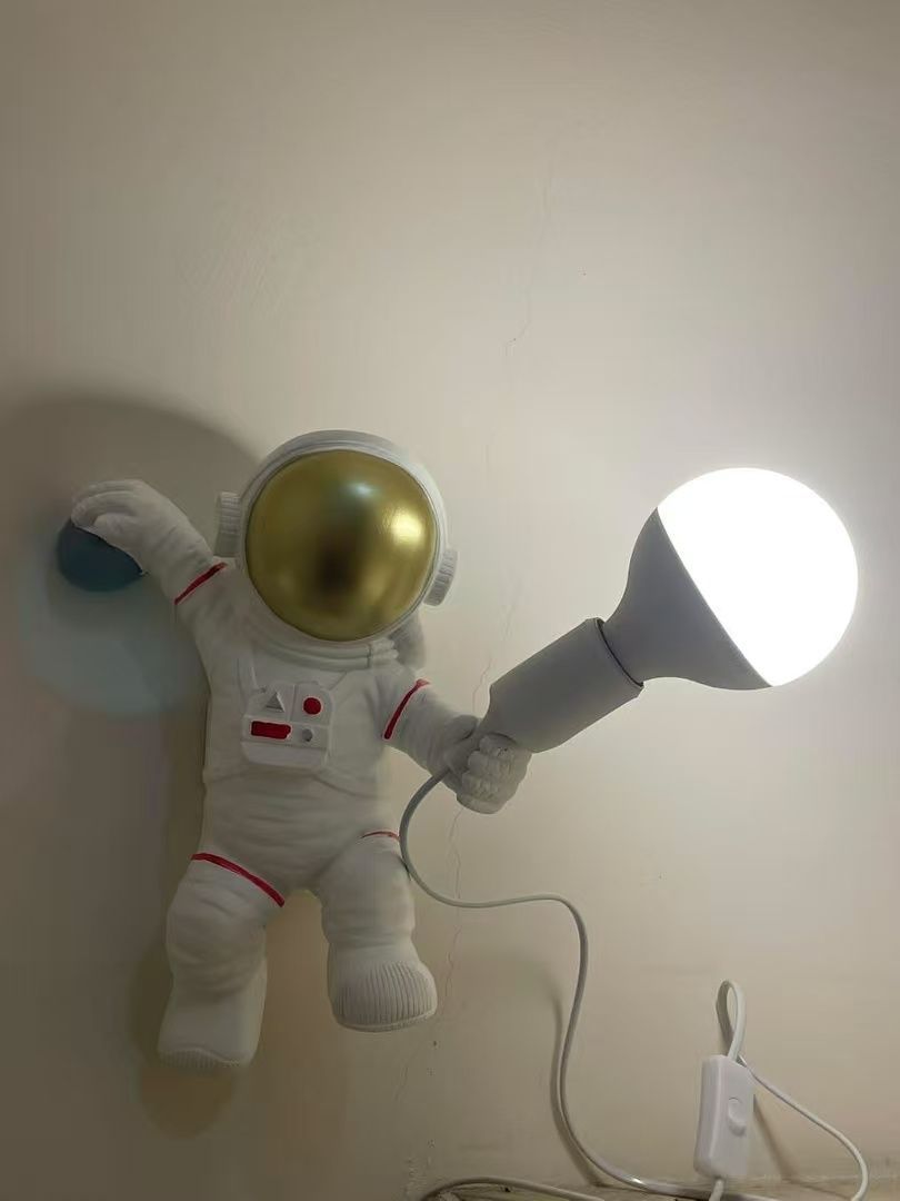 Astronaut-shaped lamp on wall; holds a lit, white bulb. White spacesuit, gold helmet.