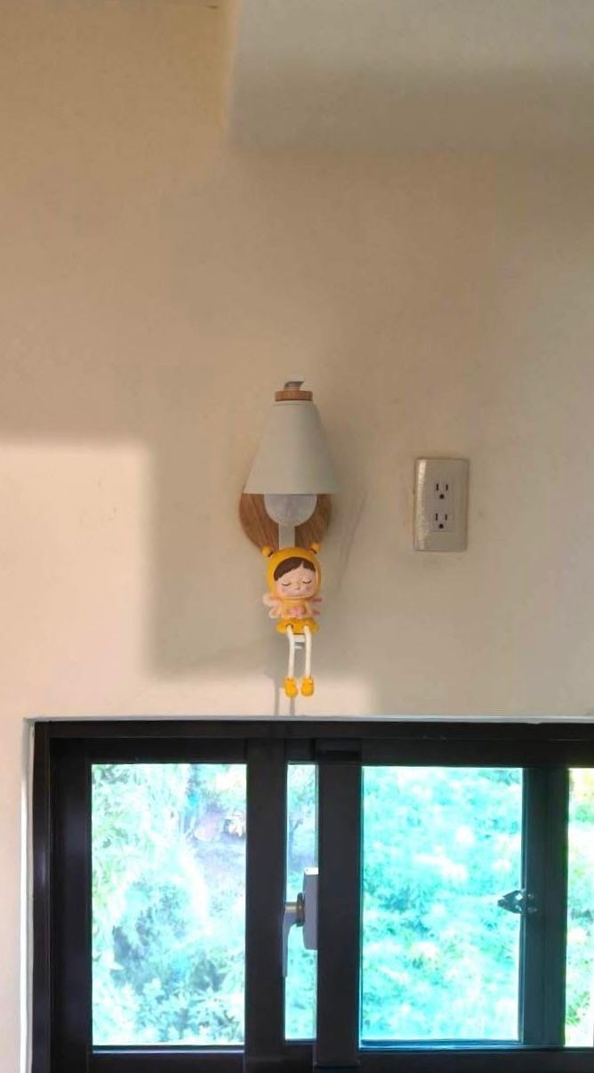 Wall lamp with cartoon figure below a lampshade, beside an electrical outlet and above a window.