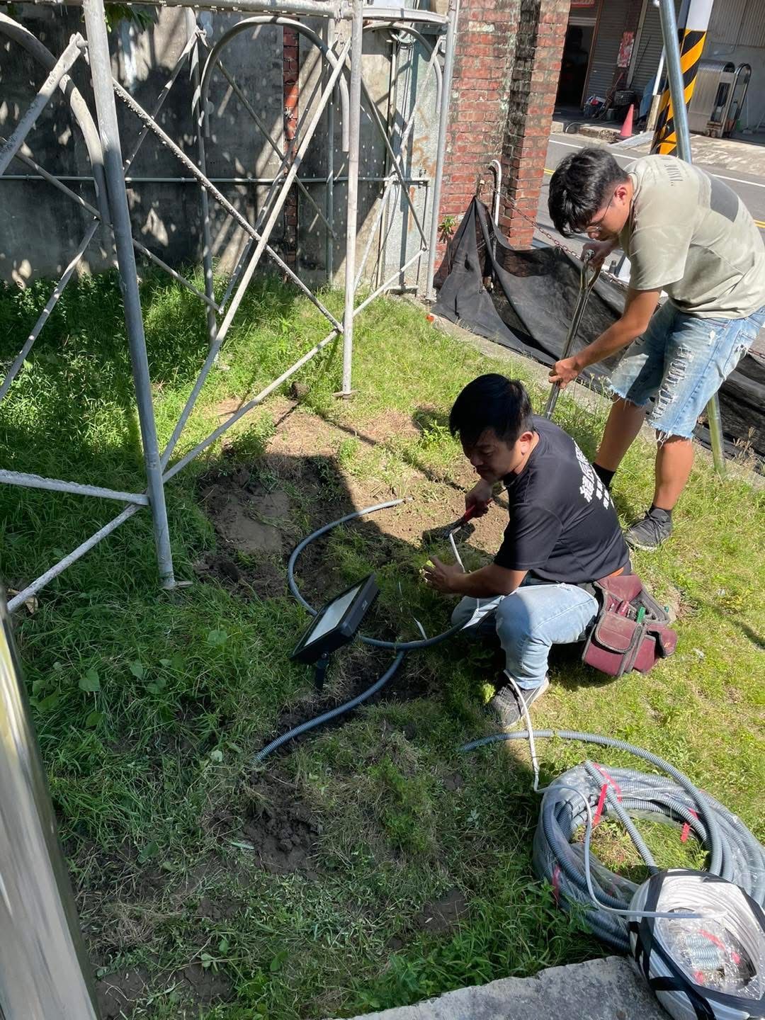 Two people working with wires near a metal structure and brick building. They are outdoors on grass.