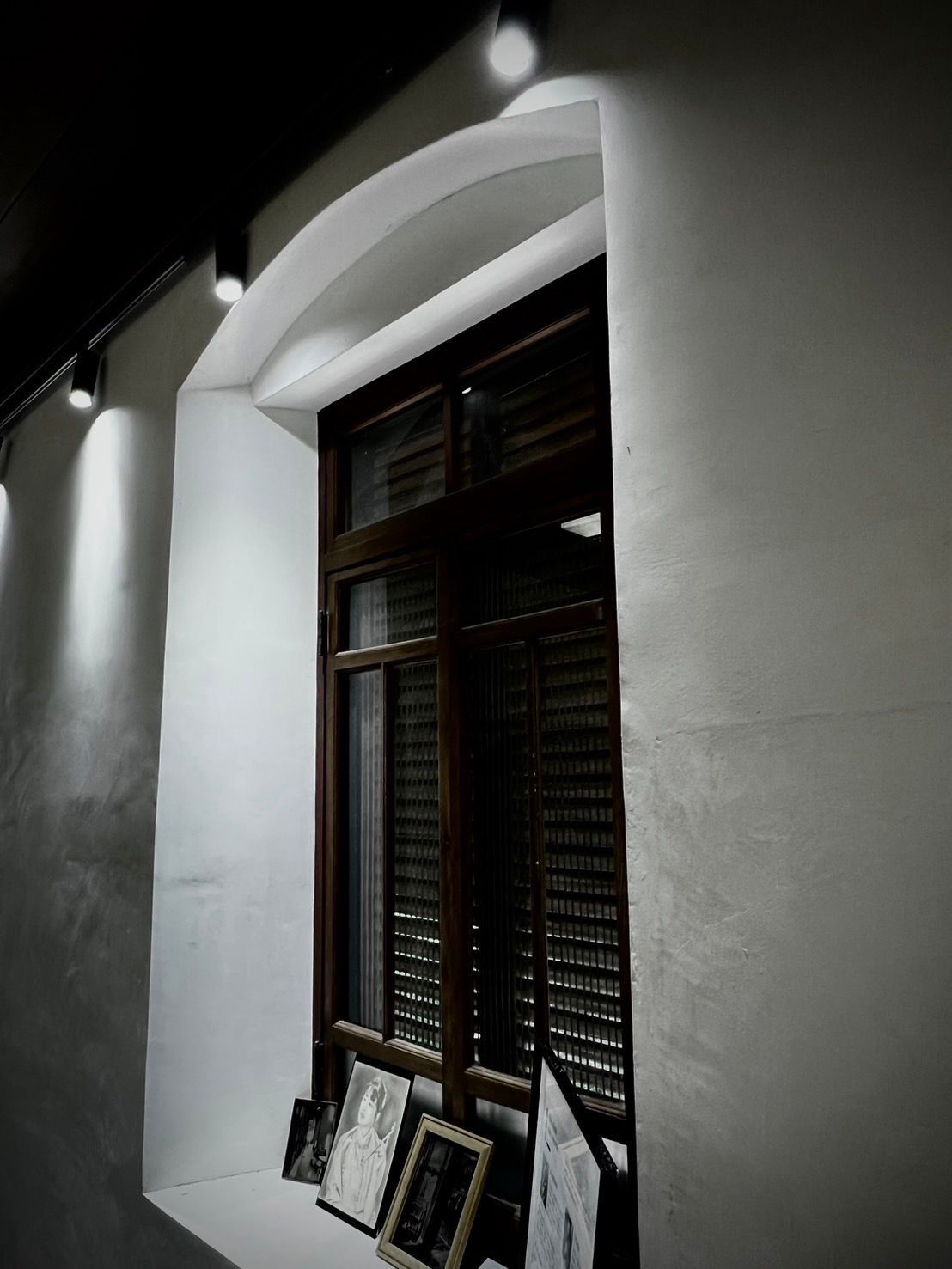 Dark interior with arched window, spotlighting wooden shutters, and framed images on the sill.