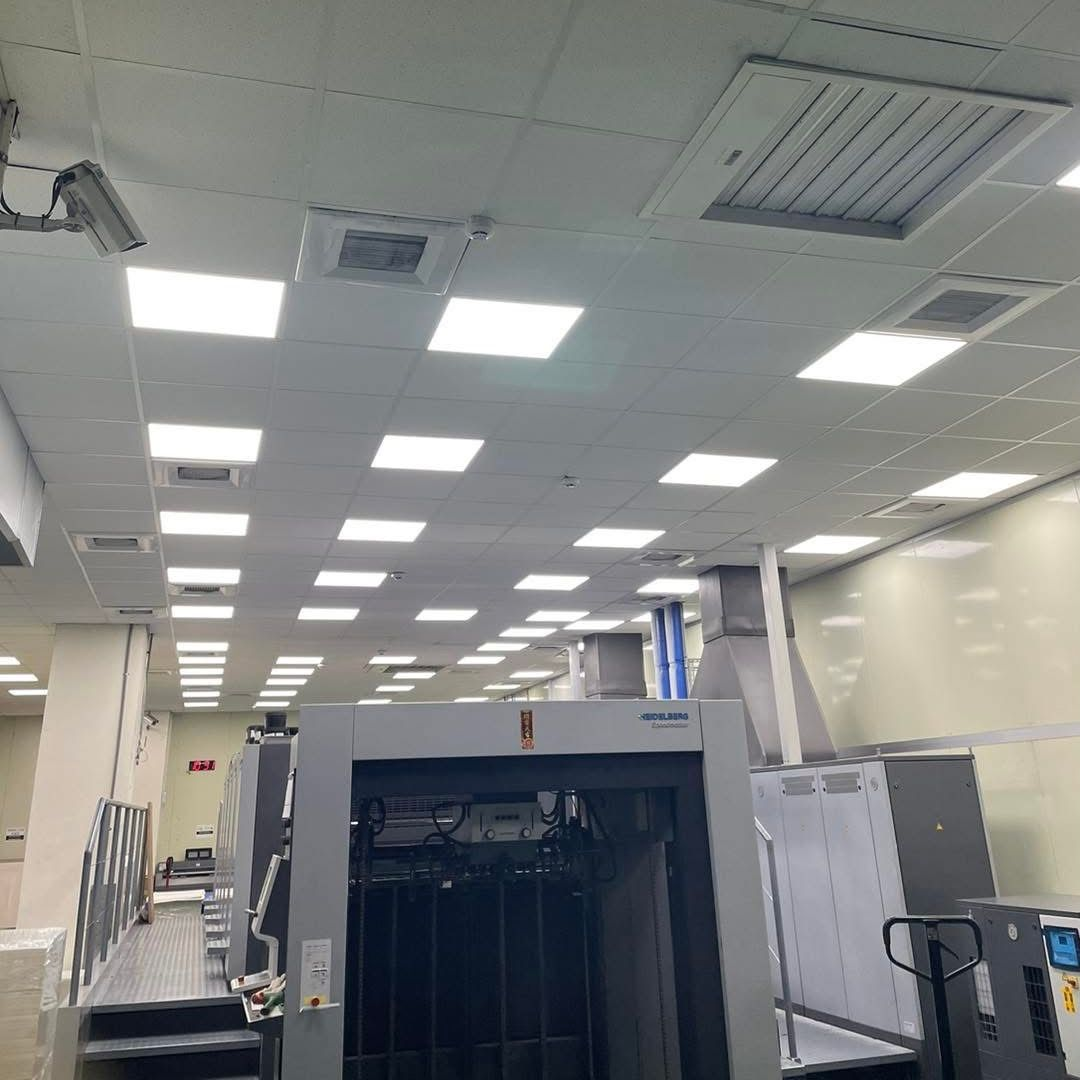 Printing press in a well-lit industrial room with numerous square ceiling lights and air vents.