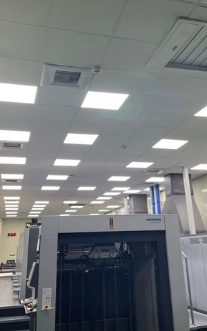 A print shop interior with large industrial printing press, fluorescent lights, and ceiling vents.