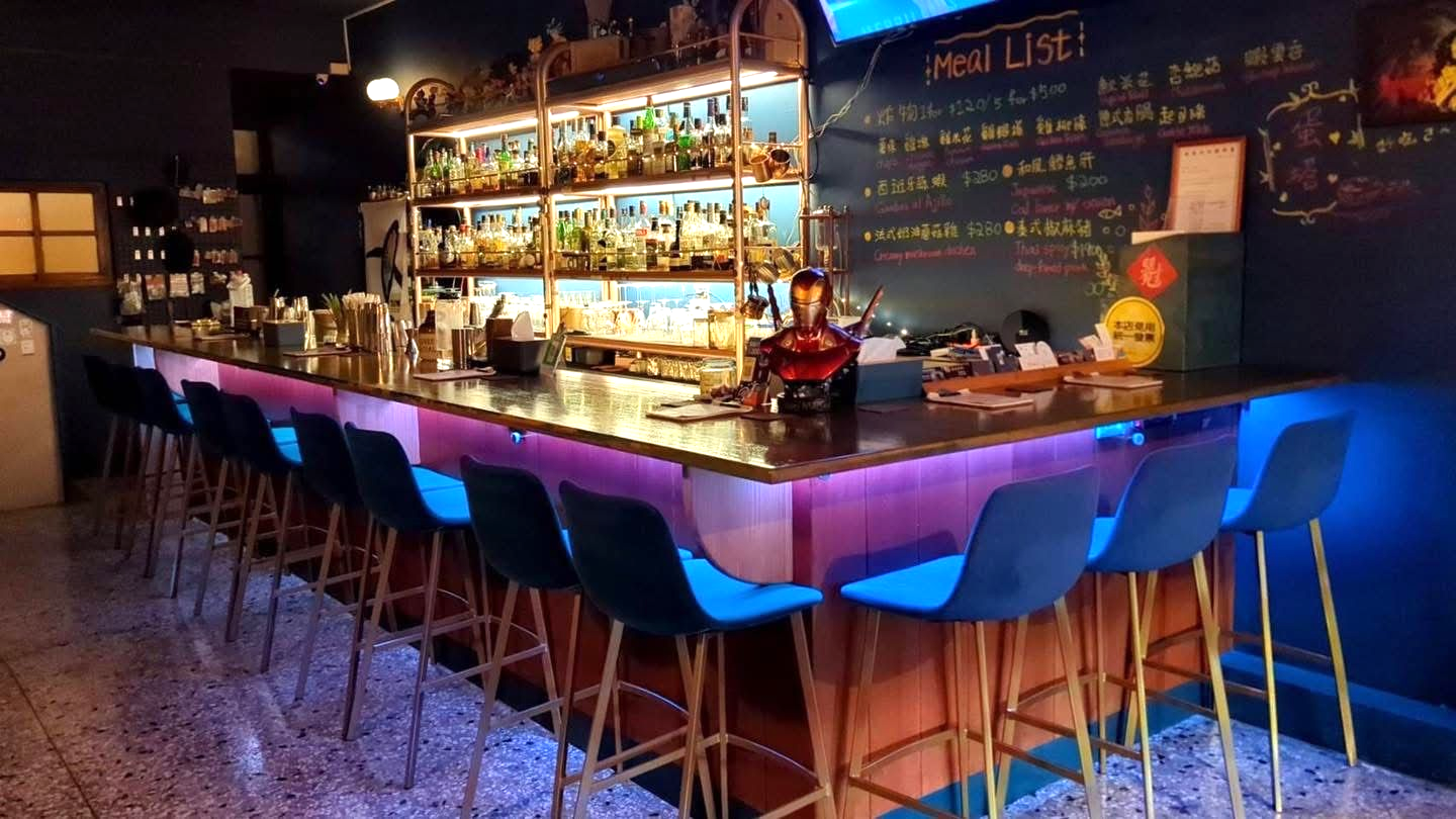 Bar interior with stools, neon lighting, shelves of alcohol, and a chalkboard menu.