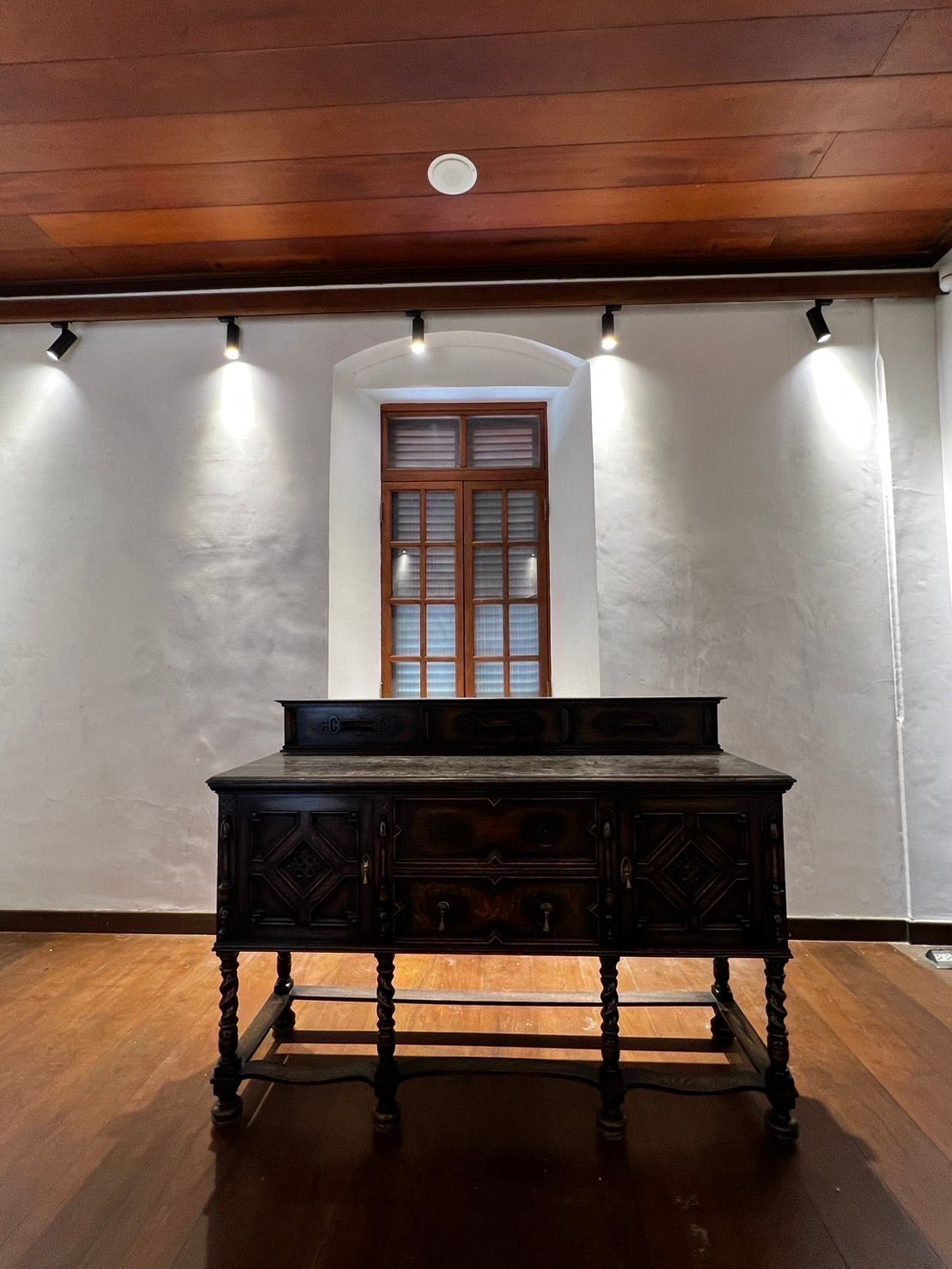 Dark wooden antique cabinet in a room with a window, white walls, and wood ceiling/floor.