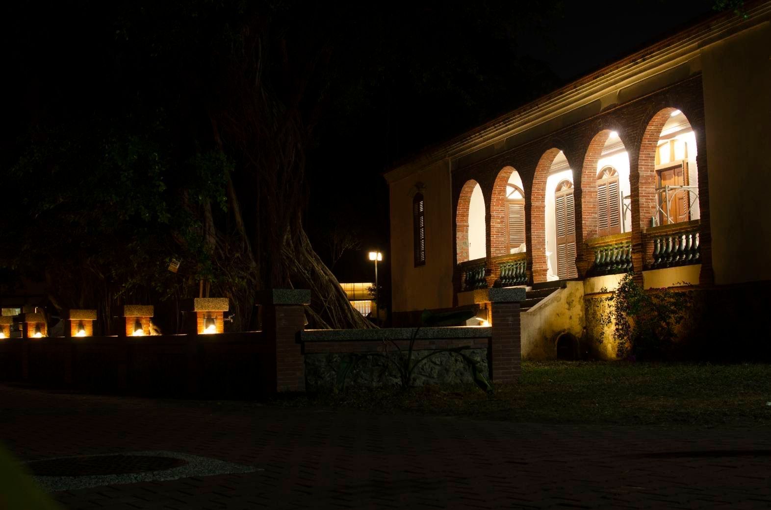 Night view of a building with arched windows and a brick patio. Illuminated letters spell out a word.