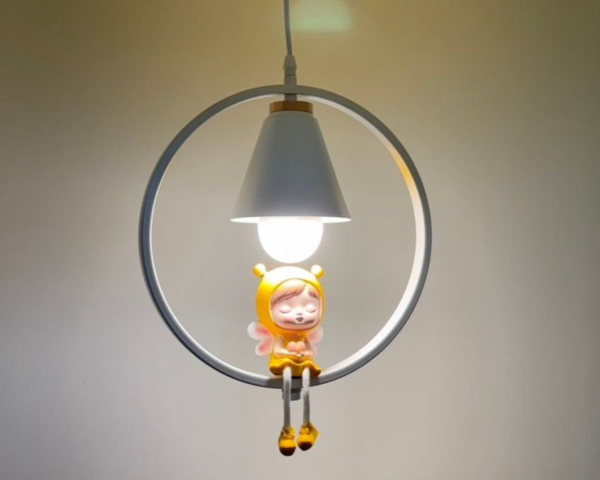 A whimsical hanging lamp with a white circular frame, a white cone shade, and a figurine of a seated fairy.