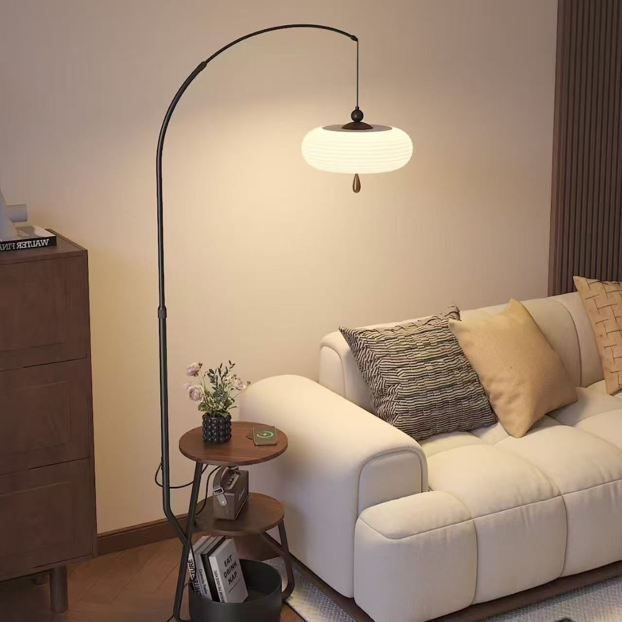 Arc floor lamp over a cream-colored couch, a side table with books, and a wooden cabinet.