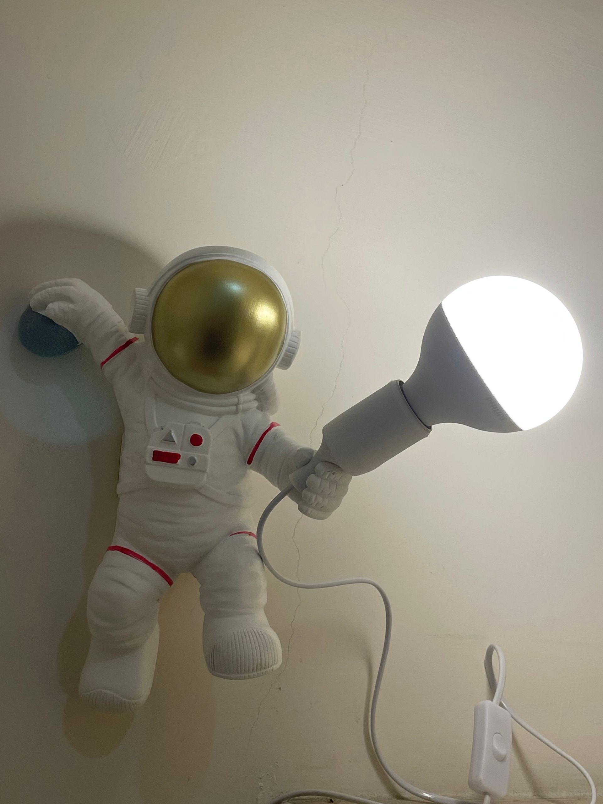 Astronaut-shaped lamp holding a large, lit lightbulb; white space suit, gold helmet, white wall.