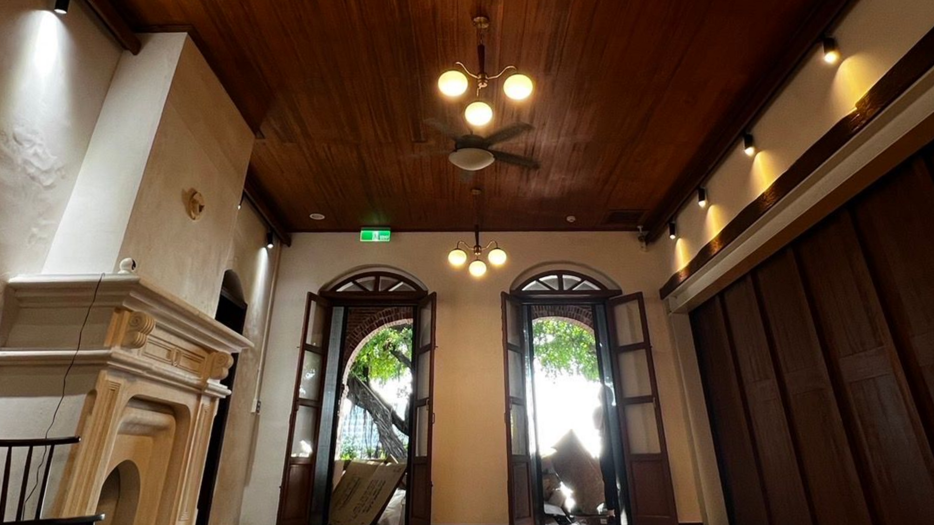Interior of a room with wooden ceiling, arched doorways, and ornate fireplace. Two hanging lights and a ceiling fan are visible.