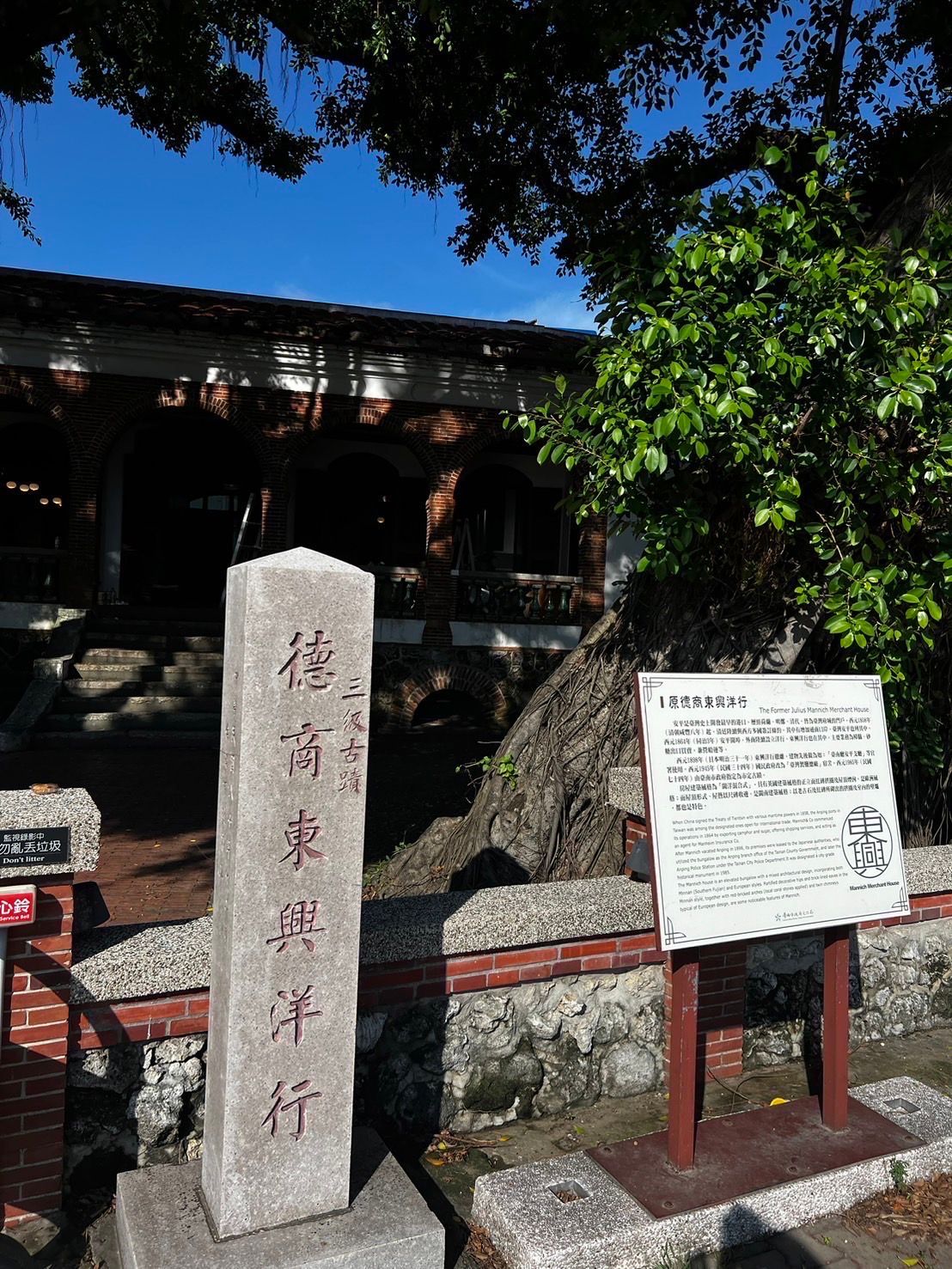Stone marker and sign outside a brick building, under a tree, clear blue sky.