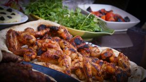 A platter of grilled chicken wings served on flatbread, accompanied by fresh herbs, grilled vegetables, and dip.