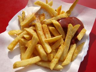 A serving of French fries on a white paper plate, topped with a dollop of mayonnaise, ketchup, and a small yellow fork.