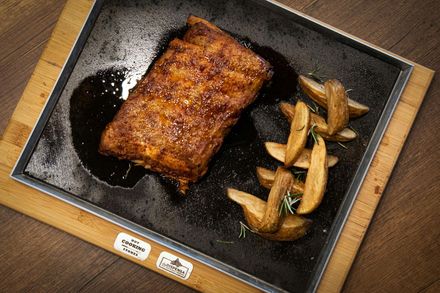 A rack of glazed ribs served on a dark sizzling platter with a side of potato wedges on a wooden board.