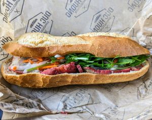 A banh mi sandwich with roasted pork, pickled vegetables, and cilantro on a baguette, sitting on branded paper wrapping.