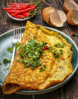 A golden-brown omelet garnished with fresh cilantro on a blue plate, with red chili peppers and eggshells in the background.