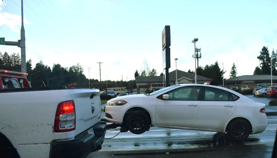 White car being towed by a white pickup truck, wheels off the ground, wet road.