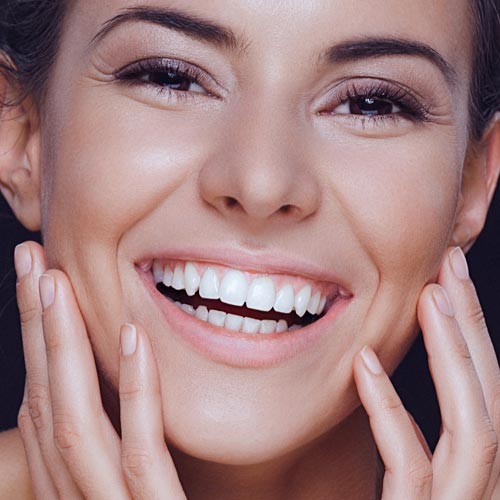 A close-up beauty portrait of a beautiful smiling young woman gently holding her clean natural face between her hands There are visible wrinkles around her eyes The woman has brown eyes healthy clean skin natural make-up and dark hair that is swept back from her face Her head is slightly tilted to the side The womans shoulders are shirtless and visible and she is isolated on a dark background Developed from RAW retouched with special care and attention small amount of grain added for best final impression
