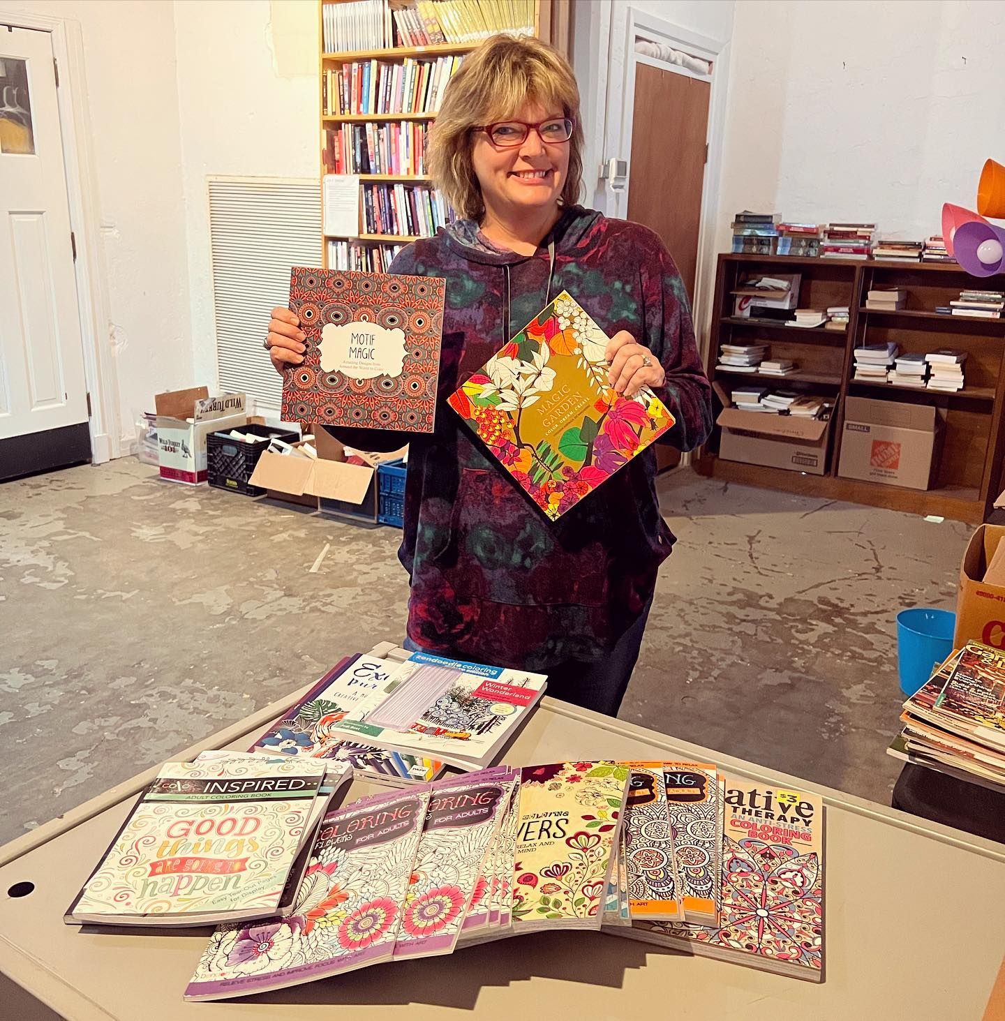 Woman holding colorful books, standing in a room with bookshelves and other books on a table.