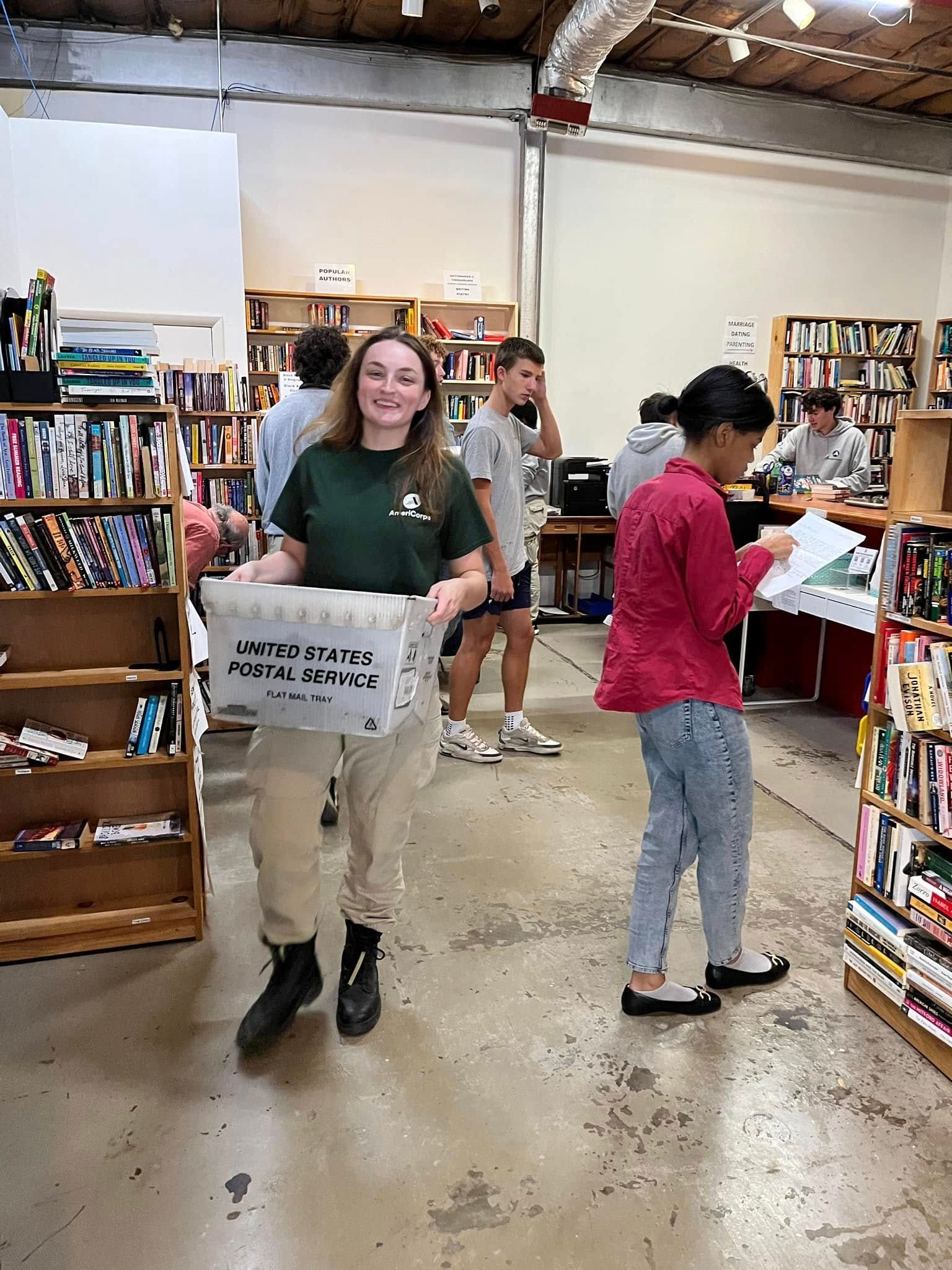 Woman smiles, carries box in a bookstore; others browse books.
