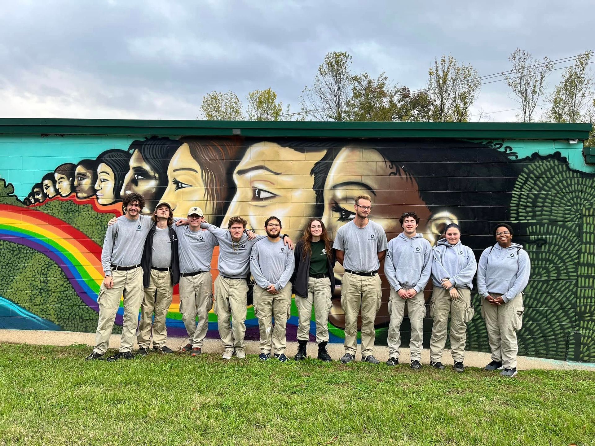 Group of people standing in front of a colorful mural featuring faces and a rainbow. They wear matching outfits.
