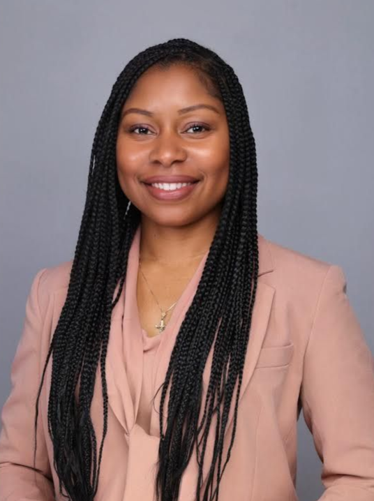 Woman with long braided hair wearing a pink blazer, smiling in front of a gray backdrop.