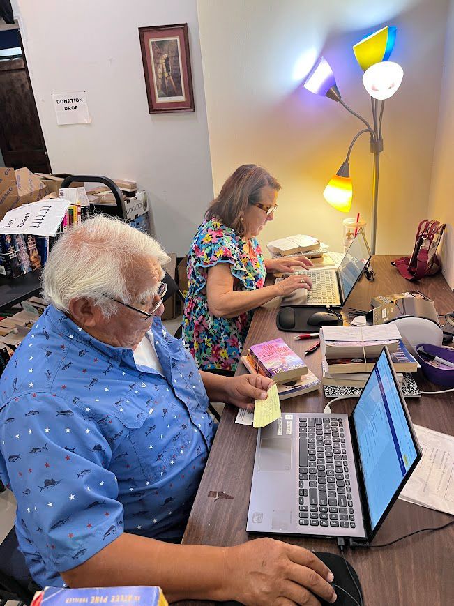Two people work on laptops at a cluttered desk under a colorful lamp.