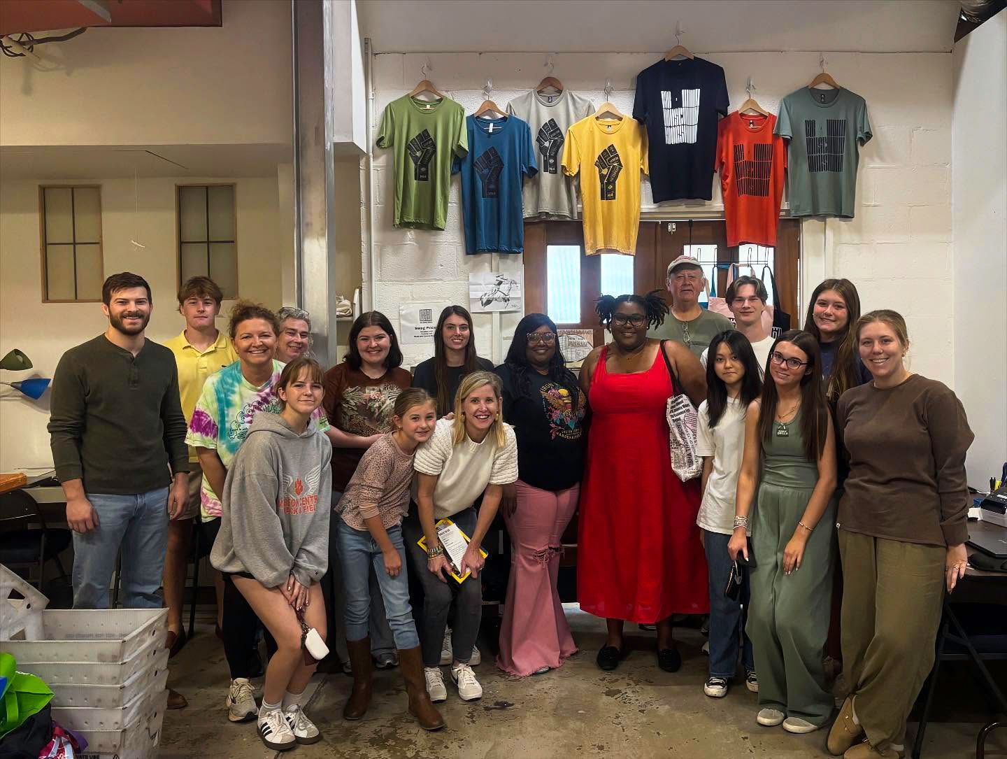 Group of people smiling, standing in front of t-shirts hanging on a wall.