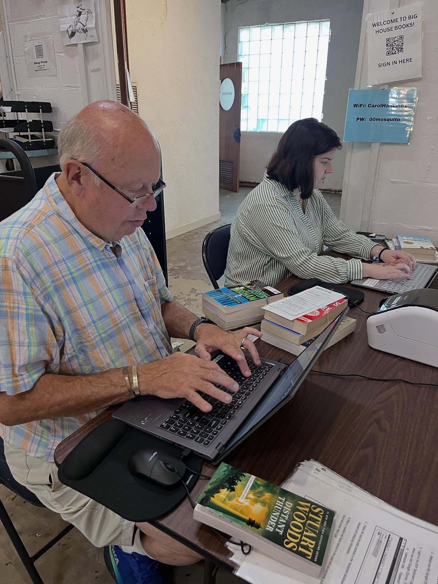 Two people work on laptops at a cluttered desk under a colorful lamp.