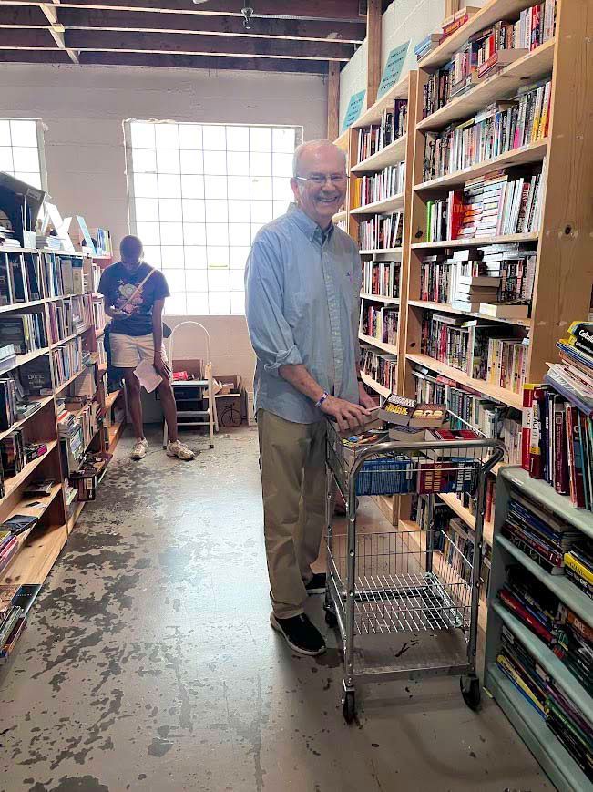 Man with cart in a bookstore, browsing shelves. A person stands in the background. Books fill shelves.