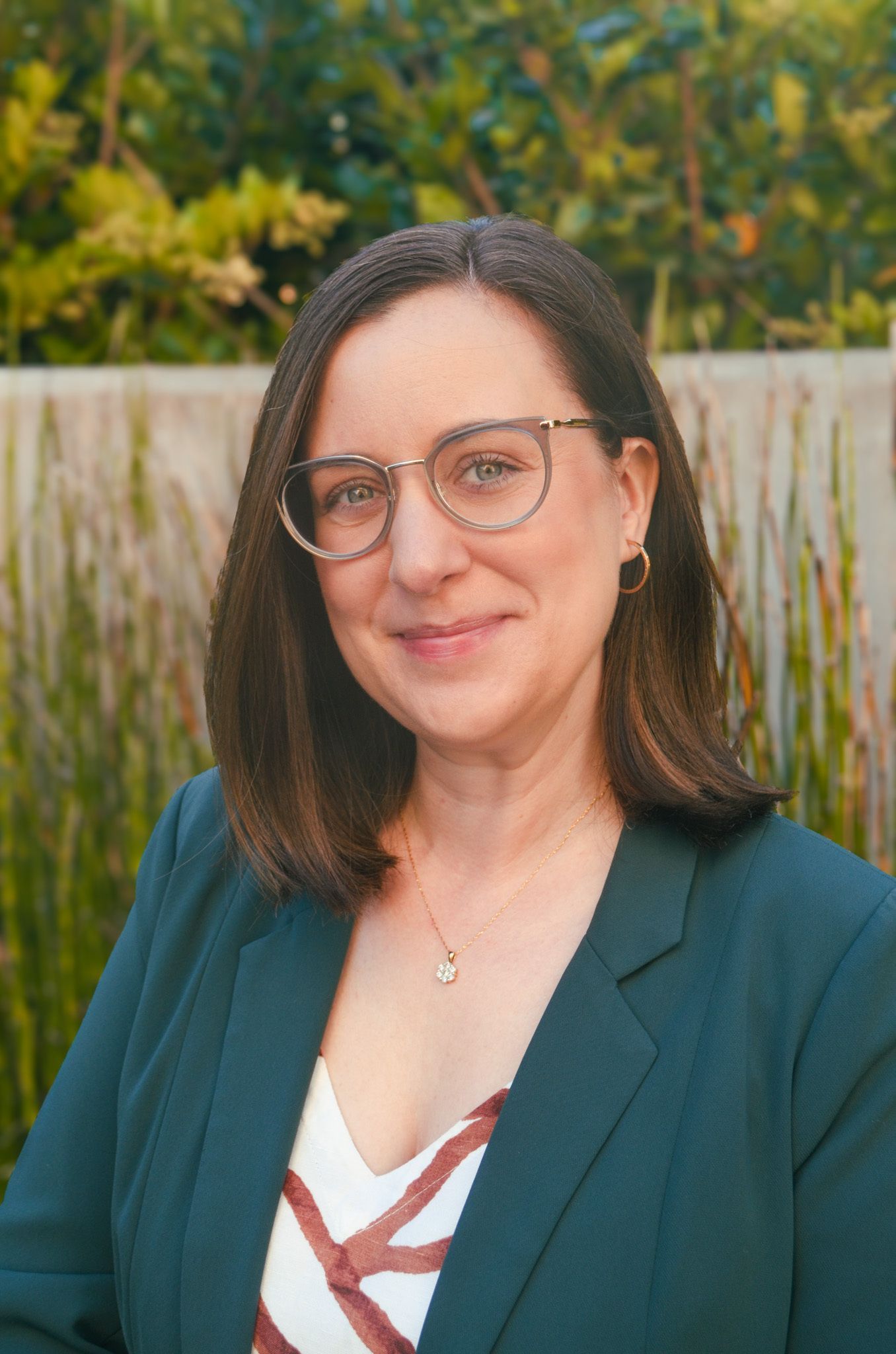 Woman with glasses and dark blazer smiles outdoors.