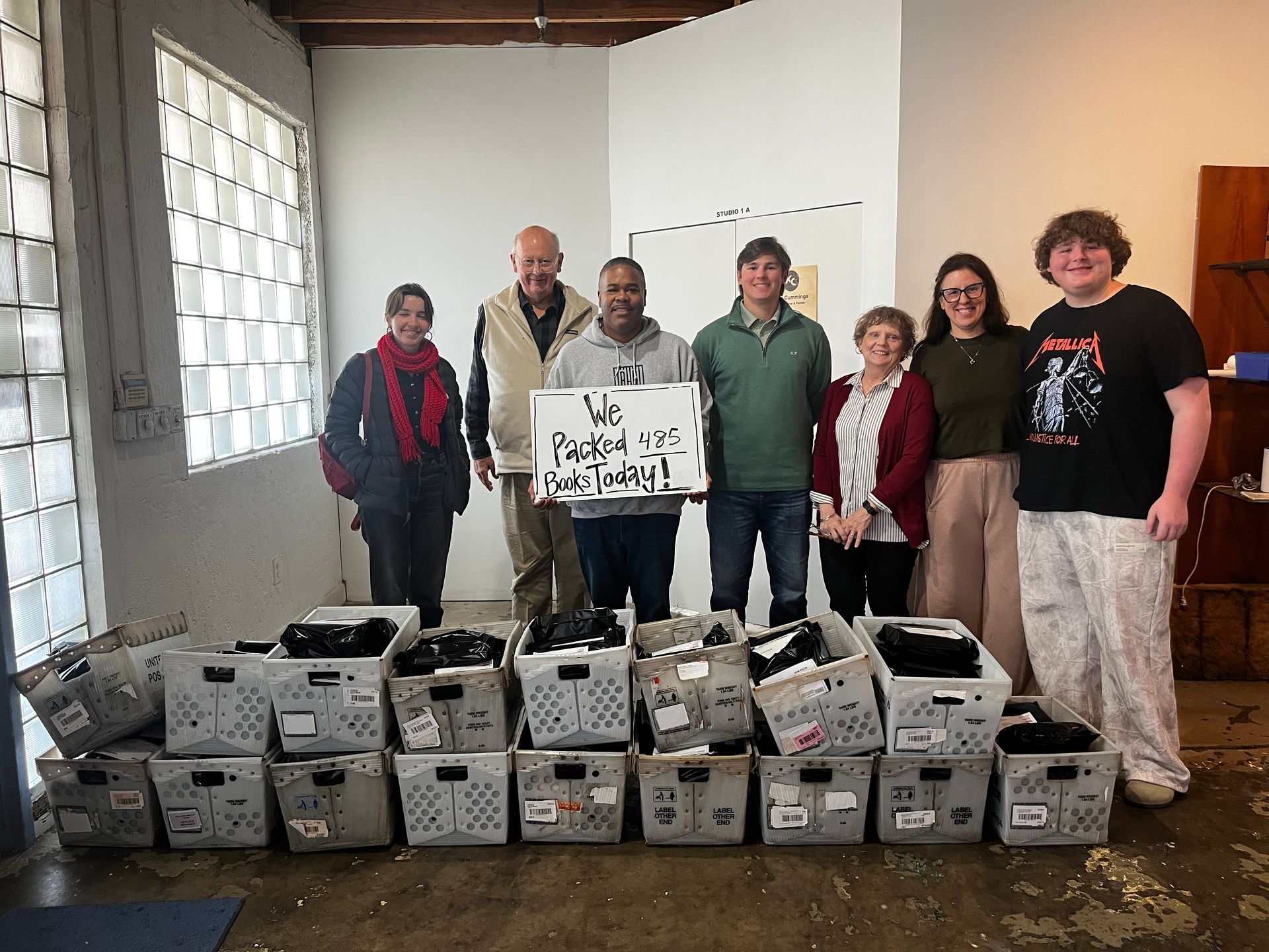 Books stacked on a wooden desk, bound with rubber bands. Shelves and a printer are in the background.