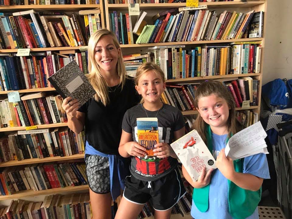 Woman and two girls smile, holding books and a notebook in a bookstore with packed bookshelves.