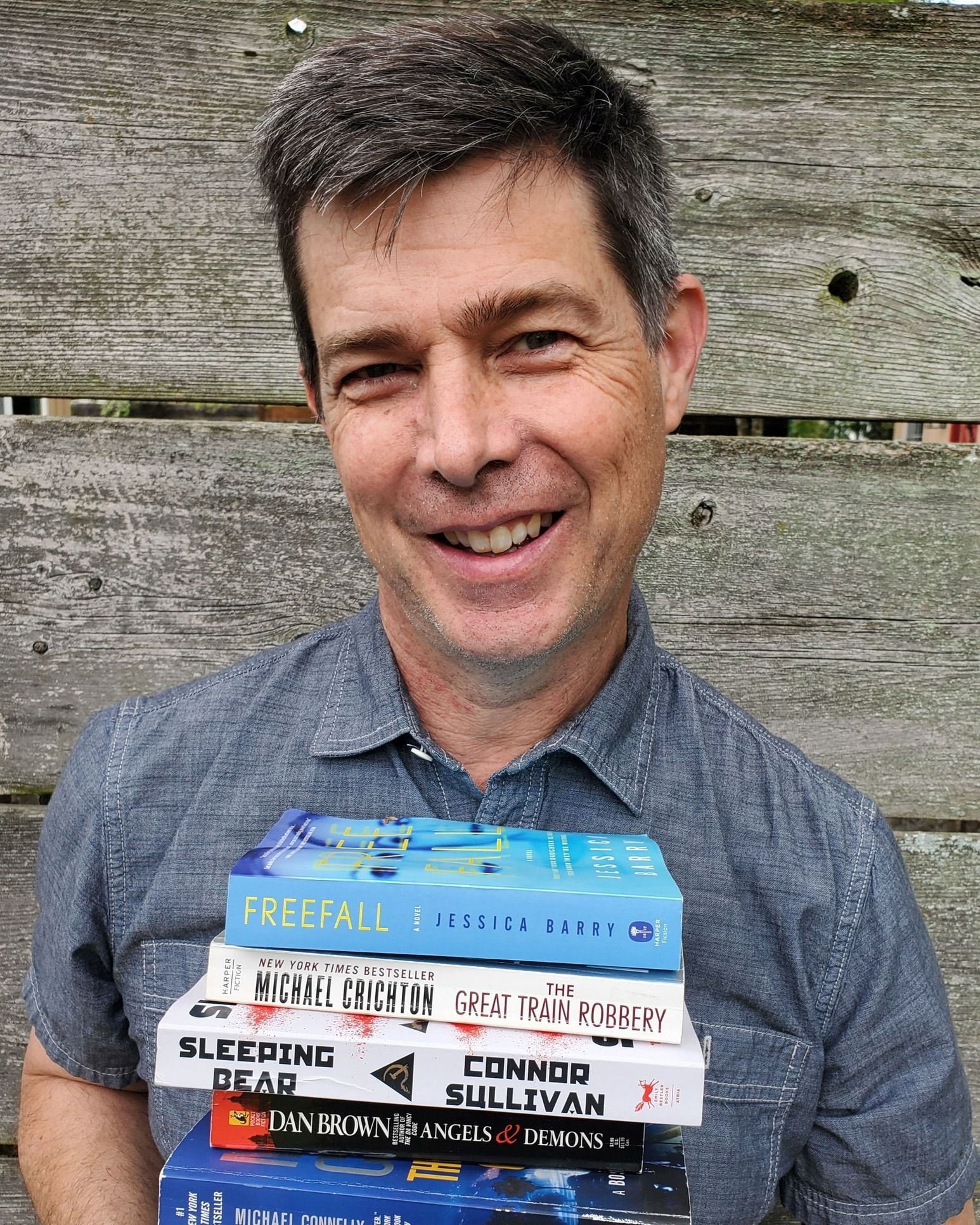 Man holding a stack of books, smiling. Gray-haired, outdoors, wooden background. Books' titles visible.