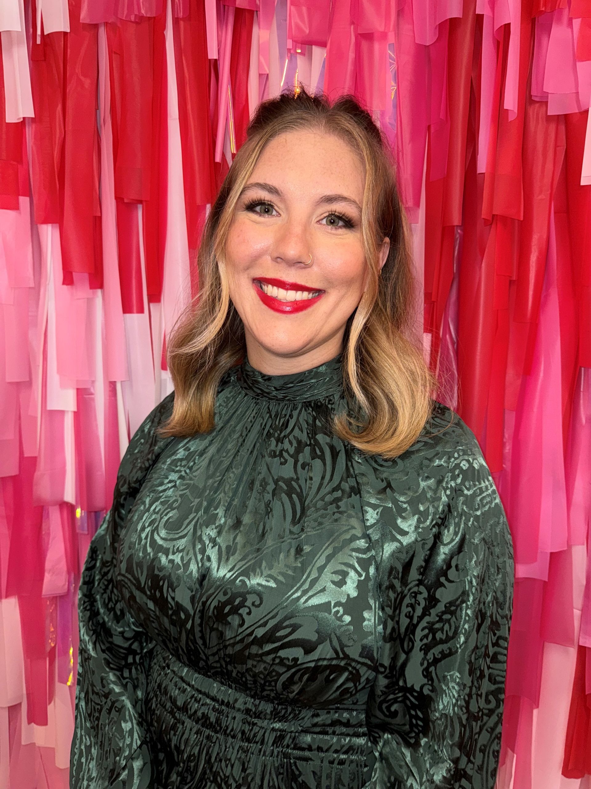 Woman in green dress smiles, red lipstick, standing before pink and red streamers.