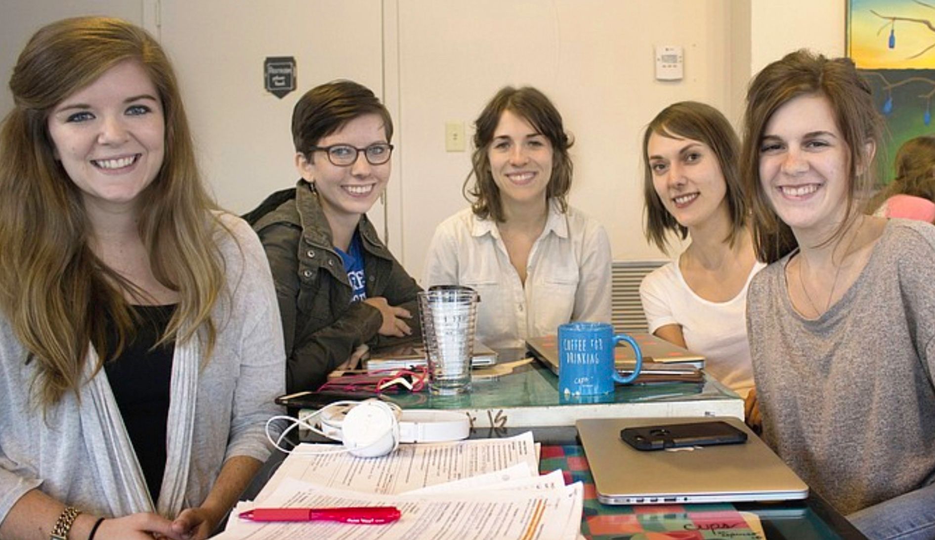 Five people sit around a table with notebooks and a laptop, smiling.