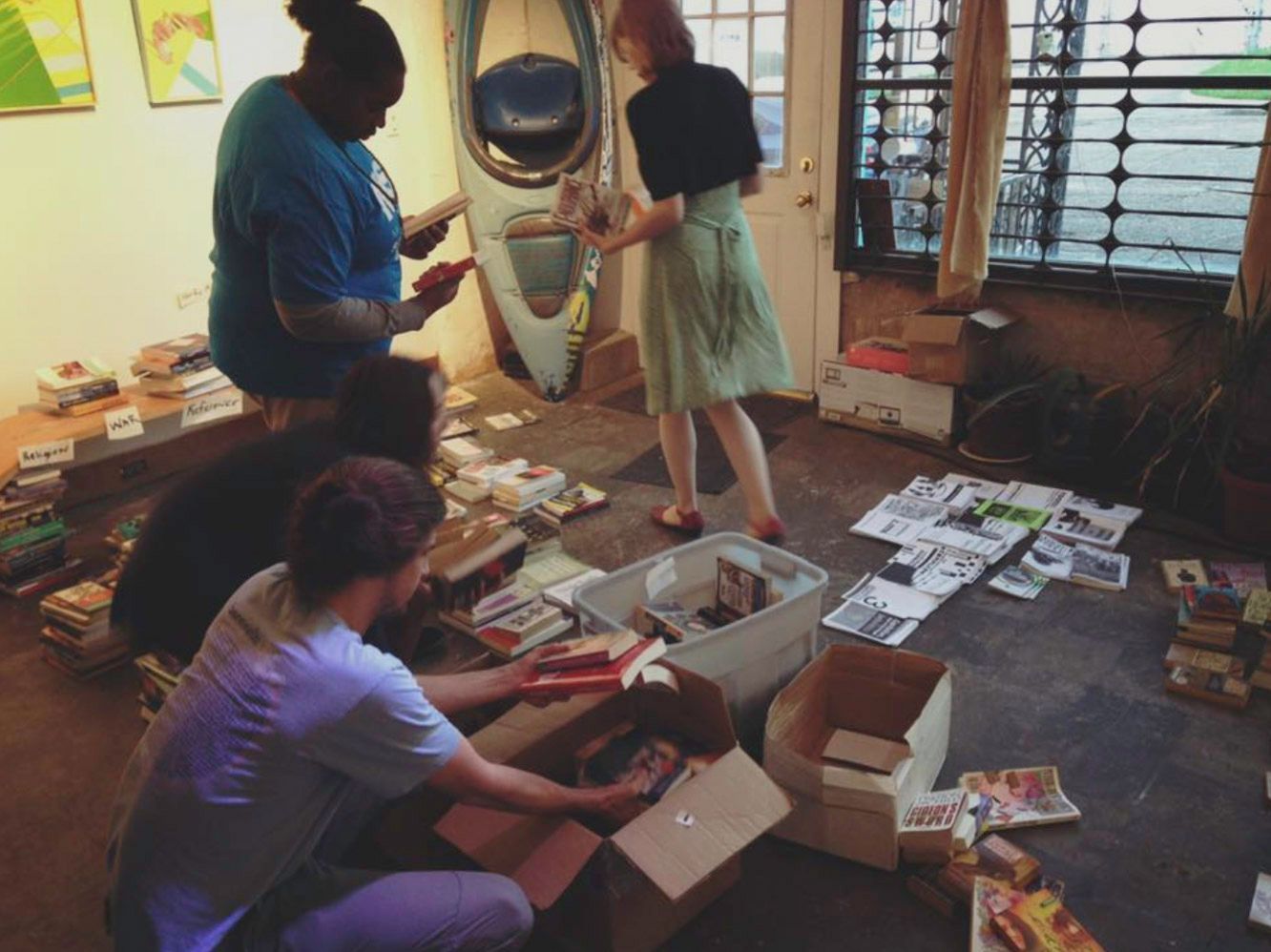 People sorting through boxes of papers and items inside a building with a kayak on the wall.