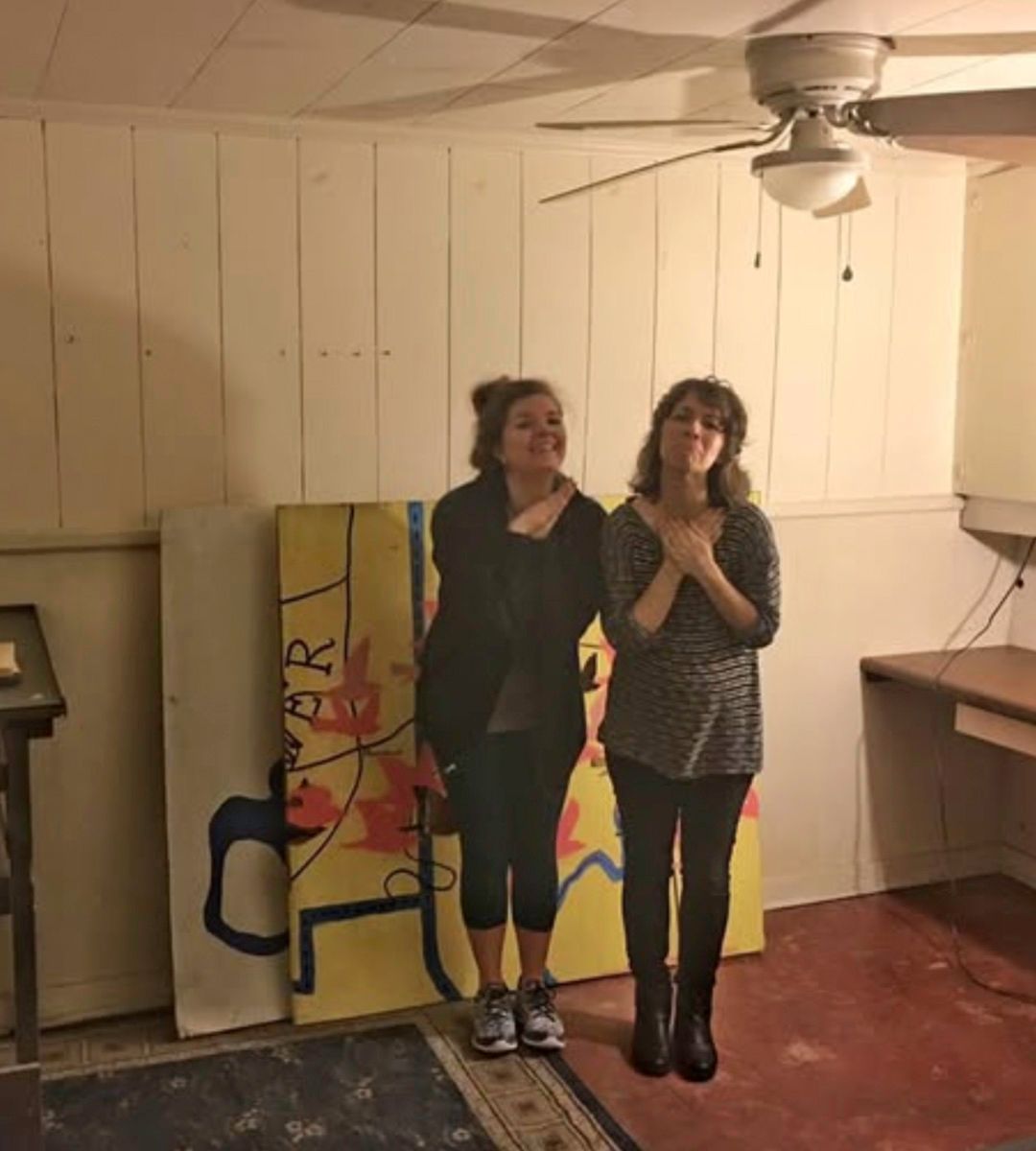 Two women in a basement smile, standing near a large, colorful painting. One has a hand on her chest.