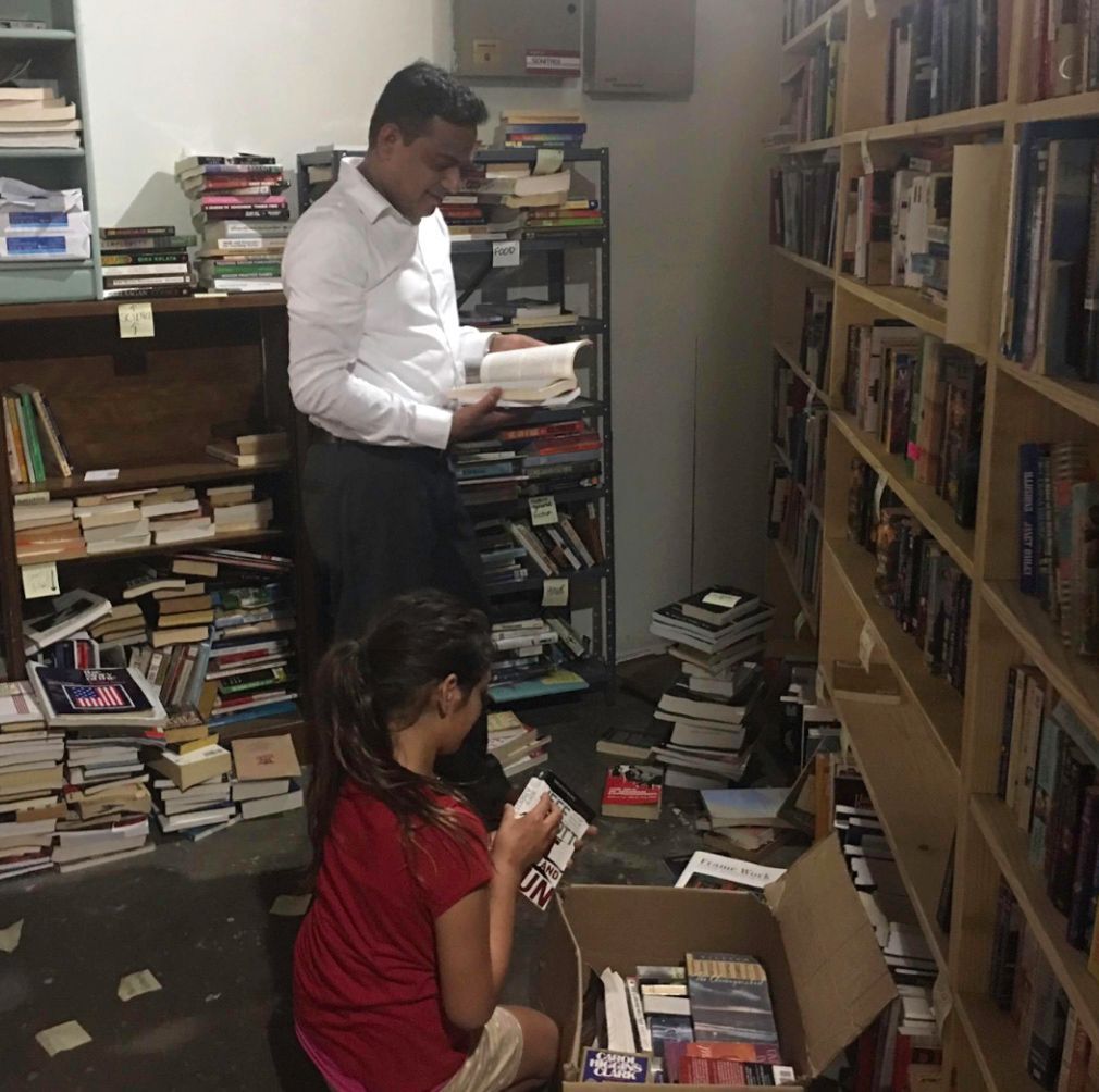 Two people organizing books in a bookstore. Shelves and boxes overflowing with books.