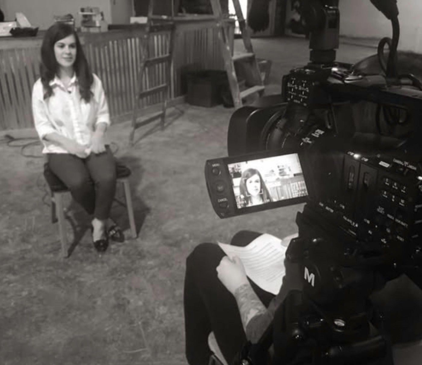 Woman being interviewed, seated on a stool. Camera filming. Indoor setting, gray tones.