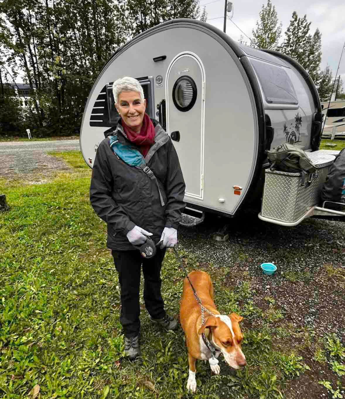 Woman and dog stand near a teardrop camper trailer. Overcast day.