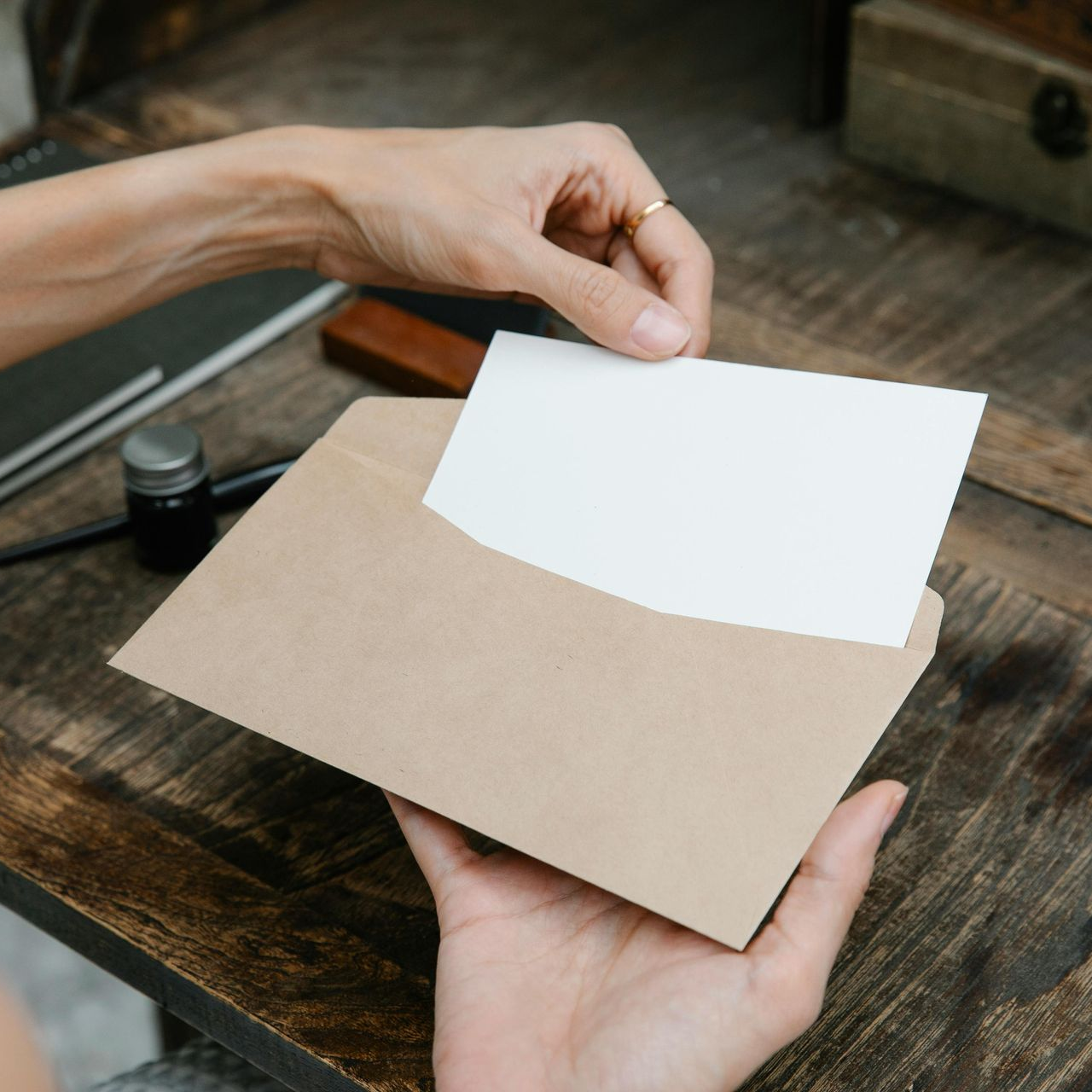 Person's hands holding a tan envelope, inserting a blank white card onto a wooden desk.