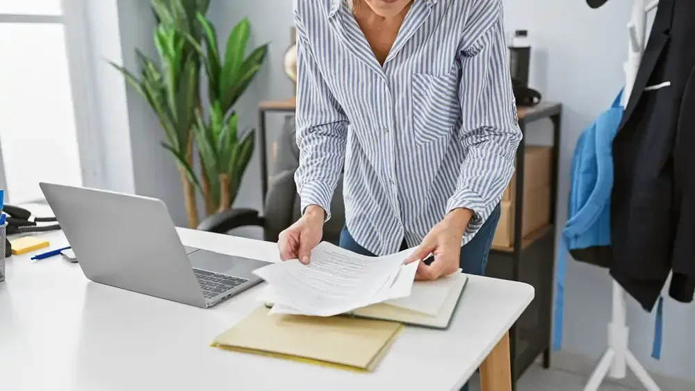 A woman is standing at a desk with a laptop and papers.