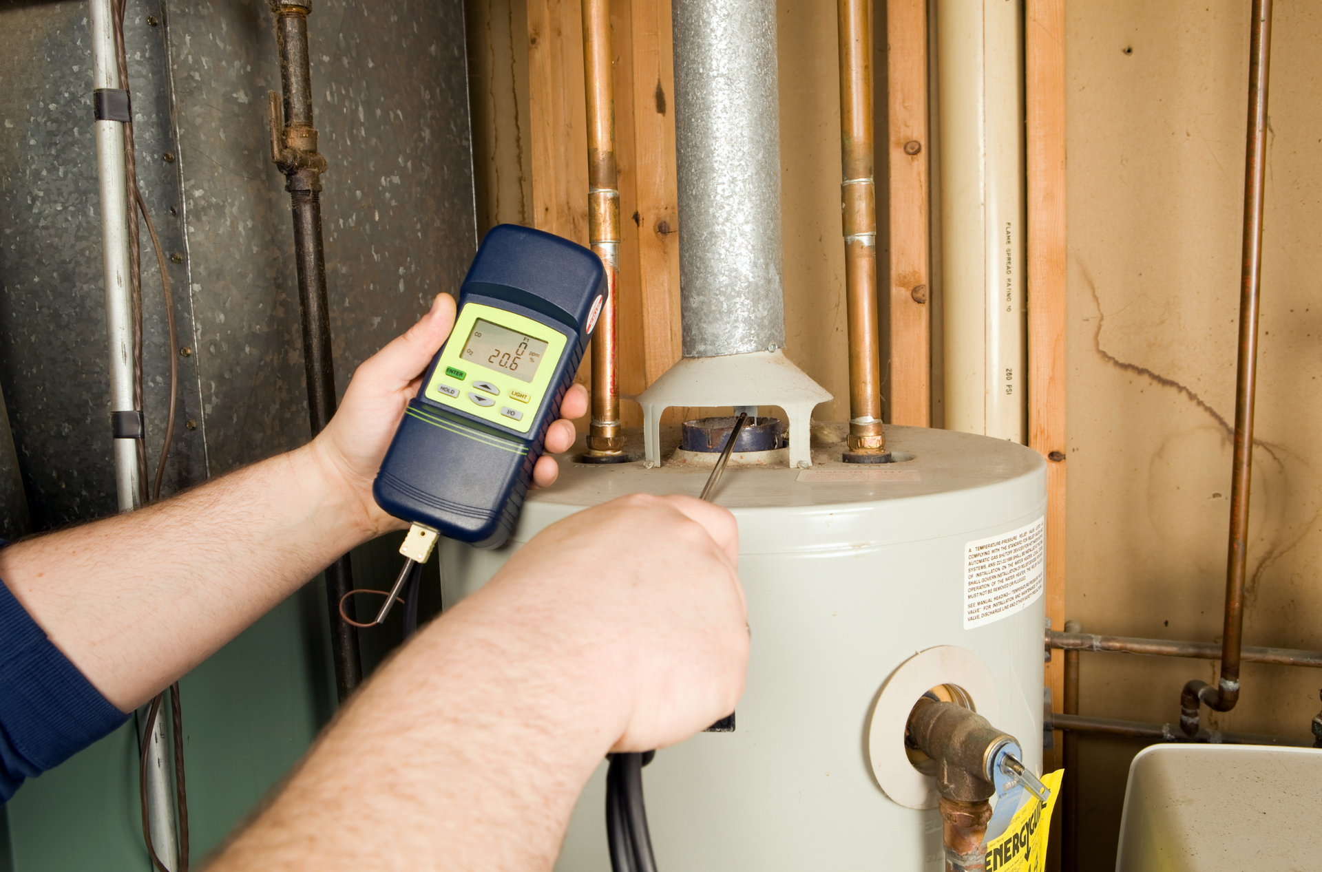 Person testing a water heater with a handheld thermometer in a utility room.
